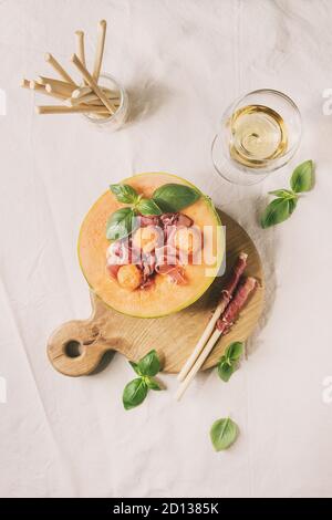 Melone e prosciutto o prosciutto insalata servita nella metà del melone Cantalupo, decorato di basilico fresco e grissini pane in legno che serve scheda sopra la linea bianca Foto Stock