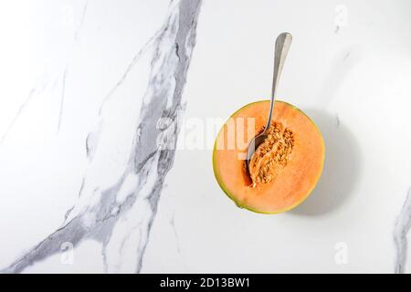 La metà di ripe organic melone melone i semi con un cucchiaio e all'interno oltre il marmo bianco sullo sfondo di texture. Laici piana, spazio Foto Stock
