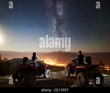 Indietro vista di uomini e ragazze che cavalcano moto quad atv sulla cima della montagna, coppia romantica godendo di una bella vista del cielo notturno pieno di stelle, Via Lattea, città luminosa sullo sfondo Foto Stock