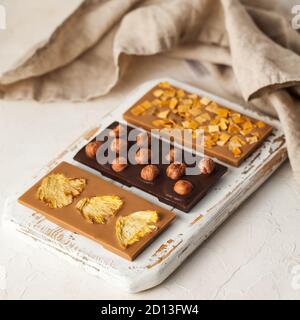 Barrette di cioccolato vegano assortite. Tre tipi di cioccolato con nocciole intere, ananas, fette di mango su una tavola di legno. Foto Stock