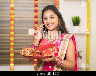 Sorridente donna indiana in abito tradizionale che tiene Karva Chauth Thali o piatto mentre occupato a parlare sul cellulare. Foto Stock