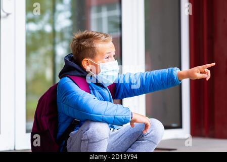 Vista laterale sorpreso indossare maschera viso giovane bambino scolaro bambino seduto su una scala vicino alla porta della scuola e puntando il dito da qualche parte Foto Stock