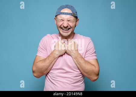 L'uomo caucasico è molto felice e in attesa di una piacevole sorpresa. Spinse i pugni al petto, chiuse gli occhi e sorrise ampiamente. Foto Stock