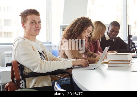 Ritratto di un gruppo multietnico di studenti che usano computer portatili mentre studiano all'università, con un ragazzo allegro che usa la sedia a rotelle guardando la fotocamera Foto Stock