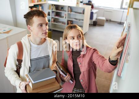 Ritratto ad alto angolo di due giovani uomo e donna che prendono i libri fuori scaffale nella biblioteca della scuola, spazio di copia Foto Stock