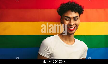 Ritratto di un bel gay che indossa make up contro bandiera di orgoglio. Sesso fluido maschio con bastone rosso e orecchino guardando via e sorridendo. Foto Stock