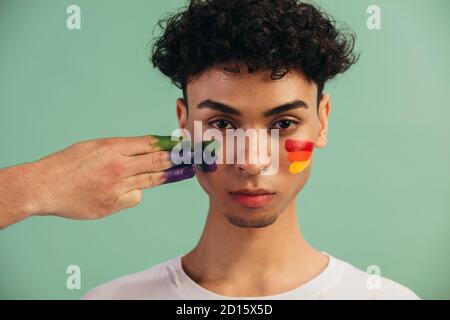 Closeup di una mano pittura LGTB bandiera colori sul viso dell'uomo. Uomo transgender con bandiera gay di orgoglio dipinta sulle sue guance. Foto Stock