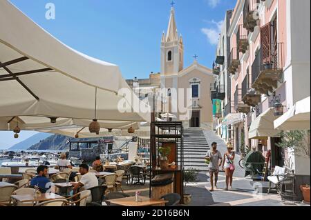 Italia, Sicilia, Isole Eolie, Lipari, coppia in outfit estivo passando di fronte alla chiesa e una terrazza caffè di Marina corta, il porto turistico di Lipar Foto Stock