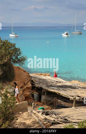 Spagna, Isole Baleari, formentera, Cala Saona, donna in abito rosso ai piedi di capannoni di barca a vela guardando ormeggi in acque turchesi Foto Stock