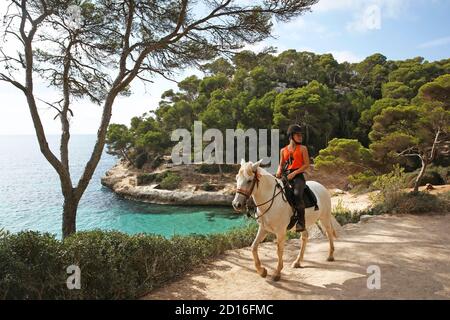 Spagna, Isole Baleari, Minorca, cala Mitjan, giovane donna a cavallo sul percorso costiero cami de Cavalls che si affaccia su un torrente selvaggio bagnata in chiaro w Foto Stock