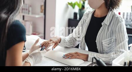 Unghie perfette e processo di manicure. Manicurista professionista in guanti copre unghie blu con top al cliente afroamericano in maschera protettiva Foto Stock