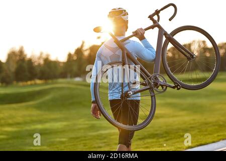 Forte atletico uomo in abbigliamento sportivo che porta la sua bicicletta e guardando il tramonto incredibile dopo aver pedalato nel parco. Stile di vita attivo e sport. Escursioni in bicicletta all'aperto Foto Stock
