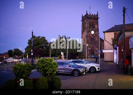 Middlewich con auto parcheggiate all'aperto, minimarket aperto fino a tardi La serata con la chiesa di San Michele sullo sfondo Foto Stock