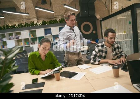 Bel capo maschio o dirigente caucasico in piedi nell'ufficio moderno e discutere di nuovo progetto con due giovani colleghi diversi seduti alla scrivania e lavorando. Lavoro di squadra. Uomini d'affari Foto Stock