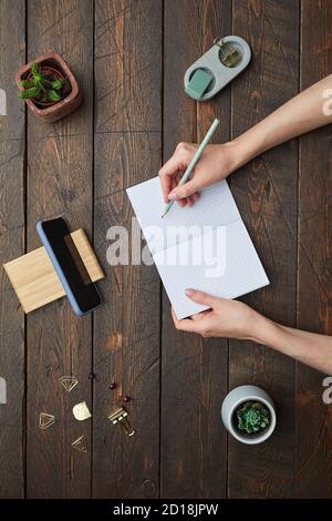 Minimo sopra la vista piatto di mani femminili che scrivono nel pianificatore sopra lo sfondo testurizzato di lavoro di legno, spazio di copia Foto Stock