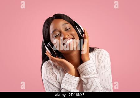 Ritratto di una bella donna afro-americana che indossa cuffie e ascolta alla bella musica su sfondo rosa studio Foto Stock