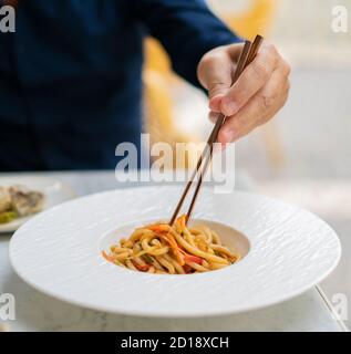 Uomo mangiare cibo vegetariano asiatico udon noodles con baby bok choy, shiitake funghi, sesamo e pepe primo piano su un piatto bianco sul tavolo. Vie frontale Foto Stock