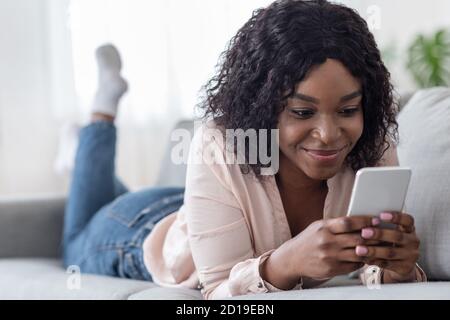 Sorridente donna africana passare il tempo con lo smartphone a casa, messaggi con gli amici Foto Stock
