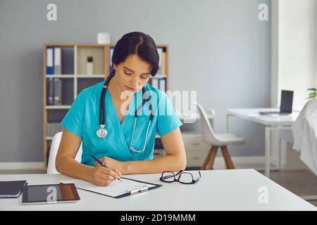 Serio medico giovane seduto alla scrivania e completare le informazioni in anamnesi medica del paziente Foto Stock