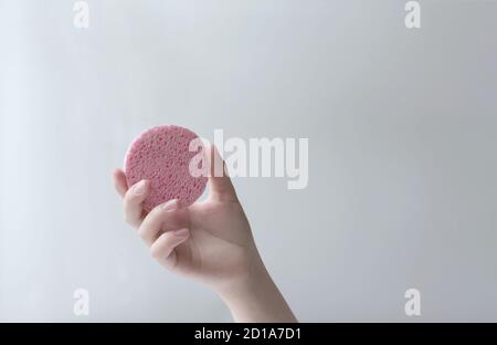 mano femminile che tiene una spugna cosmetica per la pulizia e la cura del viso. spugna rotonda rosa in mano della donna curata Foto Stock