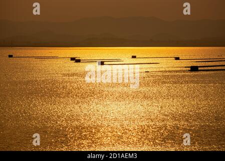 Bella natura paesaggio pesce allevamento in gabbie sull'acqua superficie che riflette la luce gialla dorata della luce solare durante il tramonto e la montagna Foto Stock