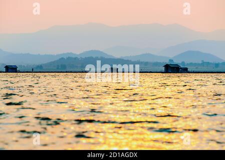 Bella natura paesaggio silhouette pescatore e gabbie di allevamento di pesce, luce del sole dorata che si riflette sulla superficie dell'acqua al tramonto, pesca locale rurale Foto Stock