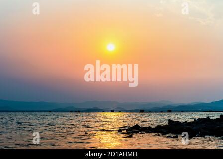 Bella natura paesaggio giallo sole nel lago cielo arancione che riflette la luce del sole dorata sulla superficie dell'acqua al tramonto, allevamento di pesce gabbie montagna b Foto Stock