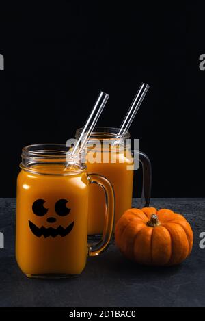 Due vaschette quadrate in vetro con coda di zucca non alcolica e cannucce in vetro su superficie nera. Il vaso è decorato con il viso nero di Halloween. Vert Foto Stock
