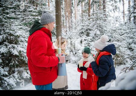 Una bella giovane coppia con bambini cammina attraverso una favolosa foresta invernale durante il nuovo anno o le vacanze di Natale. Sono riscaldati da tè caldo da fiasche thermos Foto Stock