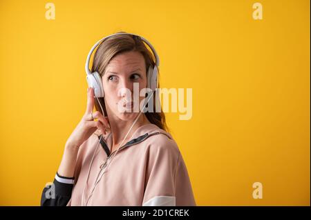Giovane donna che indossa cuffie bianche che fanno un viso d'oca. Su sfondo giallo brillante. Foto Stock