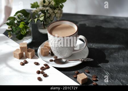 caffè bianco piatto in una tazza. cubetti di zucchero di canna e chicchi di caffè su un tavolo. fiori e luce del sole del mattino. relax Foto Stock