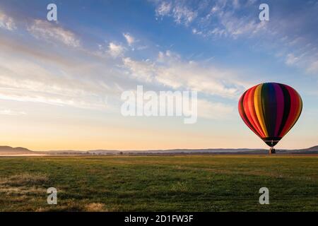 La mongolfiera in colorate strisce arcobaleno inizia a salire fattoria campo come il sole sorge blu cielo nuvoloso Foto Stock