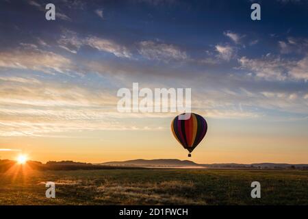 La mongolfiera in colorate strisce arcobaleno inizia a salire fattoria campo come il sole sorge blu cielo nuvoloso Foto Stock