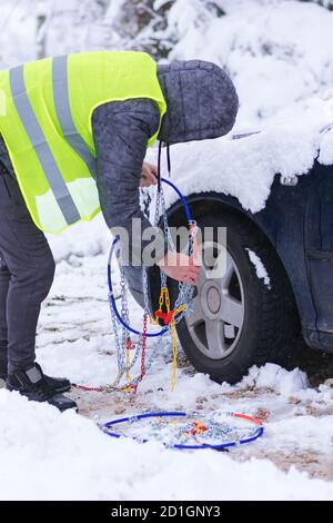 Uomo che anticipa l'auto per viaggiare durante il giorno d'inverno. Catene da neve sulle ruote dell'auto. Foto Stock