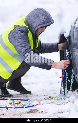 Uomo che anticipa l'auto per viaggiare durante il giorno d'inverno. Catene da neve sulle ruote dell'auto. Foto Stock