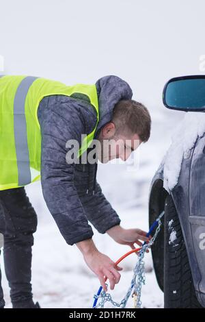 Uomo che anticipa l'auto per viaggiare durante il giorno d'inverno. Catene da neve sulle ruote dell'auto. Foto Stock