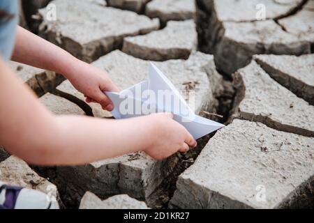 La ragazza abbassa la barca di carta sul terreno asciutto e incrinato. Crisi idrica e concetto di cambiamento climatico. Riscaldamento globale Foto Stock