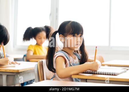 Scuola ragazza seduta a scuola scrivere nel libro con matita, studiare, istruzione, imparare. Bambini asiatici in classe. Diversità degli studenti. Foto Stock