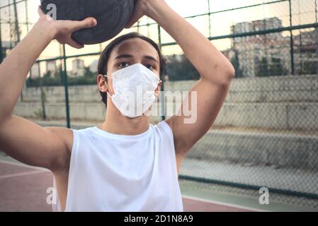 Teenager che indossa una maschera in campo di pallacanestro che si prepara a. sparare la palla tiene la palla alta sopra la testa Foto Stock