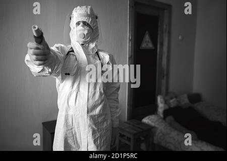 Immagine in bianco e nero del medico in tuta PPE, maschera facciale, guanti e fonendoscopio, termometro senza contatto, paziente in visita a casa durante la quarantena covid-19. Cartello di pericolo biologico sullo sportello dietro Foto Stock