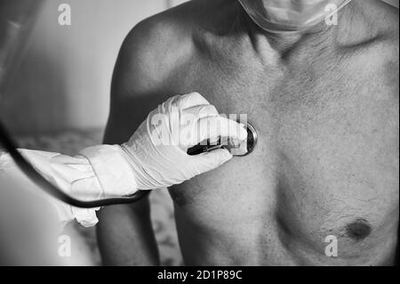 Immagine ravvicinata della mano del medico in tuta protettiva DPI che esamina il vecchio paziente di sesso maschile a casa utilizzando un fonendoscopio, controllando il petto, immagine in bianco e nero. Concetto di coronavirus Foto Stock