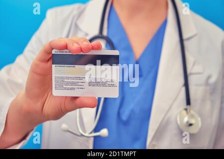 Dottore femminile che tiene una carta di credito bianca, primo piano su sfondo blu. Infermiera con stetoscopio con una carta bancaria in mano Foto Stock