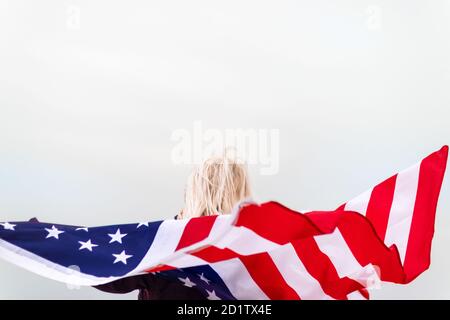 Bionda caucasica che tiene la bandiera americana appoggiata contro la schiena e guardando il mare. Bandiera USA per il giorno dell'indipendenza Foto Stock