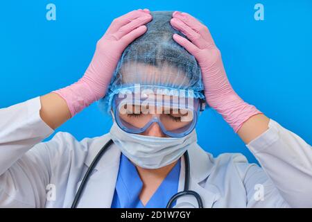 Il medico tiene le mani sopra la testa su uno sfondo blu, mal di testa. Infermiere in uniforme medica - stress e fatica dovuto coronavirus Foto Stock