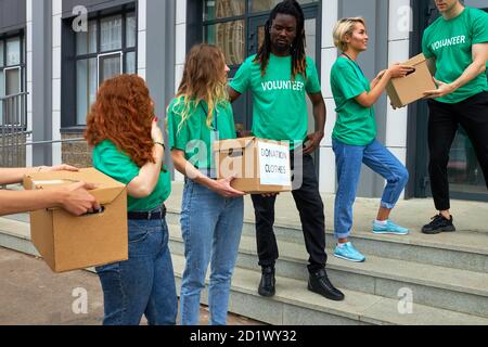 amichevole gruppo di beneficenza interrazziale impegnato in volontario, si levano in piedi all'aperto su scale trasfere scatole con cibo e vestiti, concetto di donazione Foto Stock