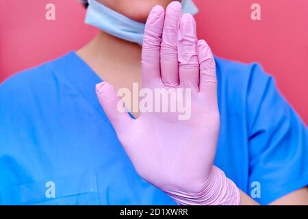 Un medico in uniforme blu su sfondo rosa mostra un gesto di arresto con la mano. Medic in guanti protettivi con un segno di arresto per pazienti con co Foto Stock