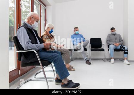 Coppia senior con maschere di fronte sedute in una sala d'attesa di un ospedale insieme ad un uomo giovane e maturo - mettere a fuoco sul vecchio uomo in primo piano Foto Stock