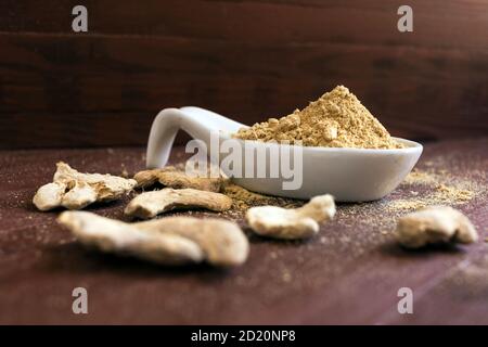 Zenzero in polvere artigianale biologica in cucchiaio di porcellana bianca e zenzero secco su sfondo marrone. Foto Stock