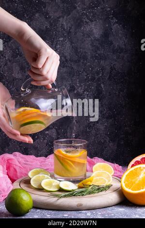 La ragazza versa la limonata di arancio, limone, lime, pompelmo e rosmarino da una caraffa di vetro in un bicchiere su un supporto di legno su sfondo scuro Foto Stock