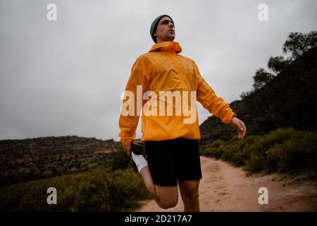 Bello giovane corridore di sentiero che allunga il muscolo della gamba prima di correre sopra sentiero di montagna in tempo nuvoloso con cespugli Foto Stock